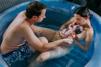 Mama und Papa begrüßen ihr nacktes Baby direkt nach der Hausgeburt im Pool in München